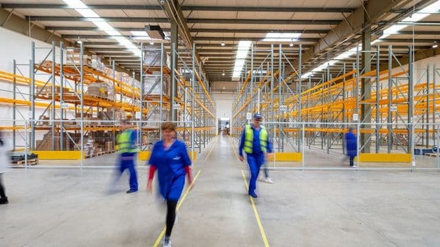 Profissionais de uniforme azul e colete caminham entre as estantes altas de um centro de distribuição moderno e iluminado