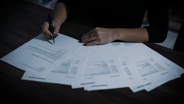 Mãos de auditora com caneta analisam um leque de documentos financeiros sobre a mesa escura