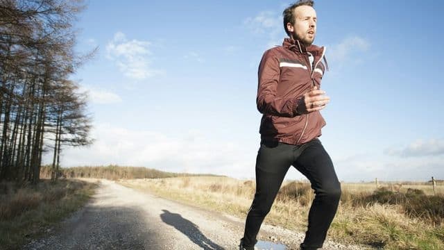 Homem corre em estrada de cascalho entre campos sob céu azul, cena de aptidão física ao ar livre, foto Jenny Hill