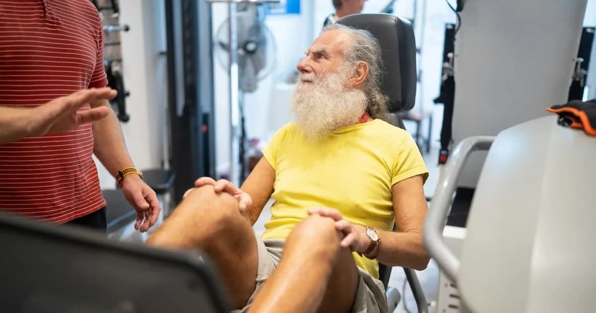 Idoso de barba branca em equipamento de reabilitação ouve a orientação do fisioterapeuta na academia terapêutica