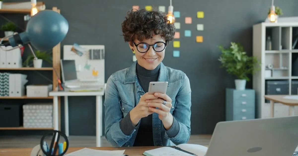 Mulher sorri enquanto mexe no celular em um escritório criativo de comunicação, com post-its coloridos ao fundo