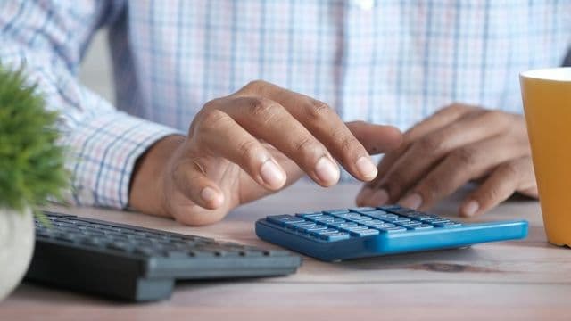 Contador digita em calculadora azul ao lado de teclado e caneca amarela sobre a mesa de trabalho