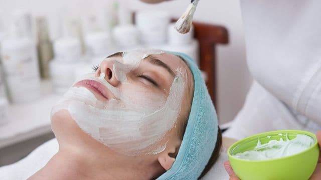 Esteticista aplica máscara facial com pincel no rosto de cliente deitada com touca de toalha, em clínica de estética com prateleira de cosméticos ao fundo