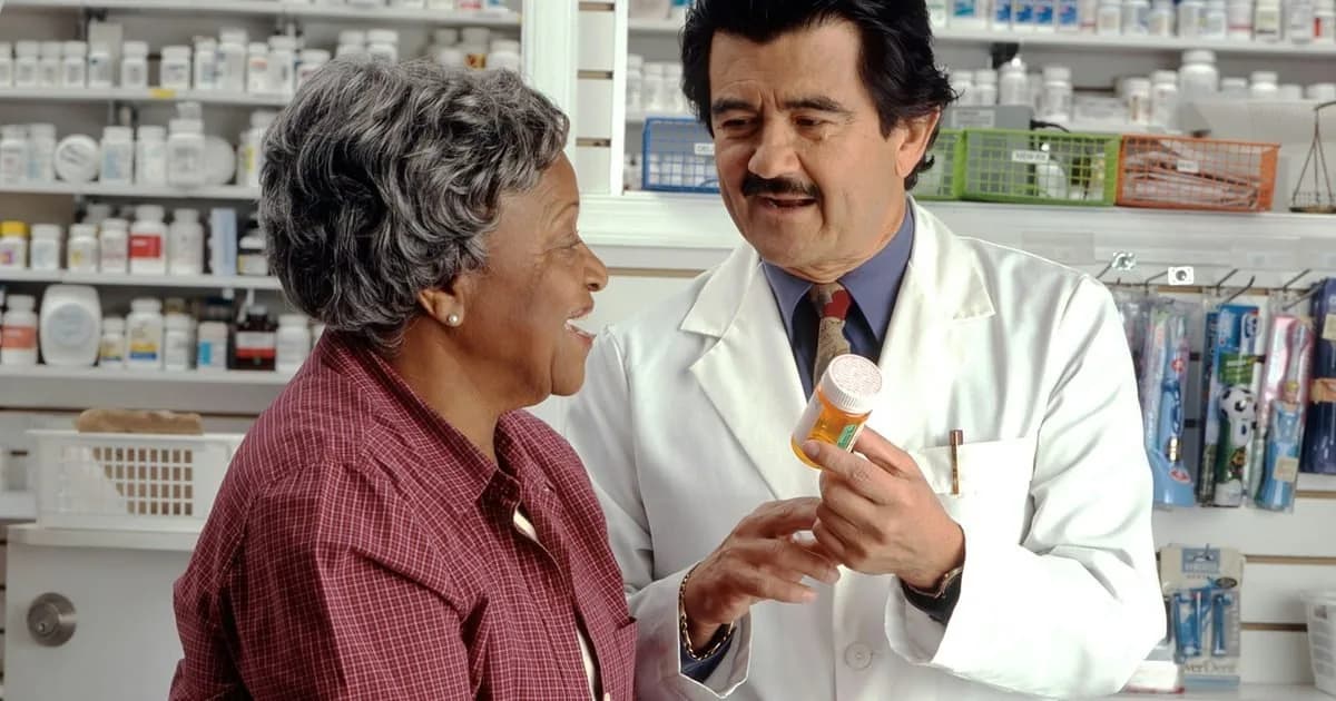 Farmacêutico de jaleco branco orienta uma cliente no balcão de uma farmácia, com prateleiras de medicamentos ao fundo