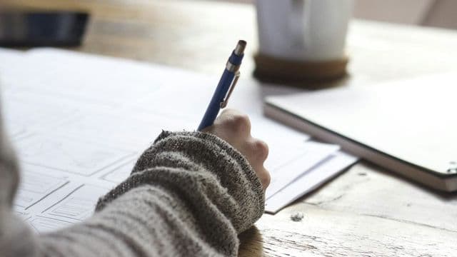 Mão escreve com caneta azul em folhas de papel sobre mesa de madeira, ao lado de caderno e caneca