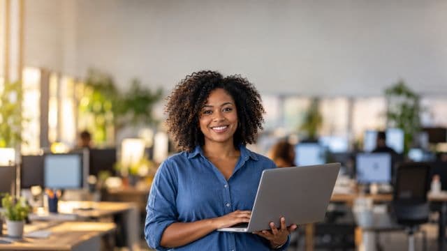 Desenvolvedora de software sorrindo, com camisa casual azul, segurando um notebook aberto, em um escritório de tecnologia moderno com monitores desfocados