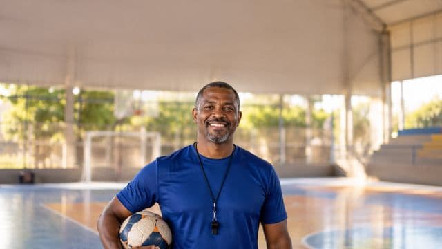 Profissional de educação física sorrindo, com camiseta esportiva azul e apito no pescoço, segurando uma bola, em uma quadra poliesportiva coberta
