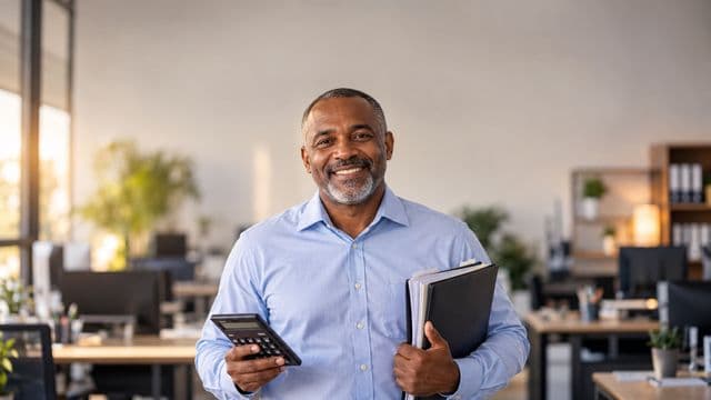 Contador sorrindo, com camisa social azul-clara, segurando uma calculadora e uma pasta de documentos, em um escritório contábil moderno