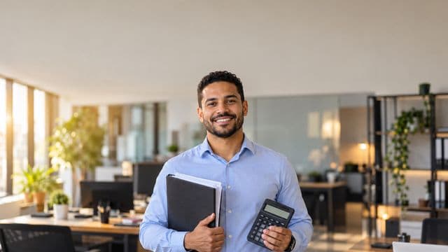 Contador sorrindo, com camisa social azul-clara, segurando uma calculadora e uma pasta de documentos, em um escritório contábil moderno
