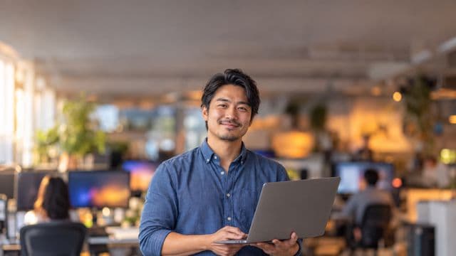 Desenvolvedor de software sorrindo, com camisa casual azul, segurando um notebook aberto, em um escritório de tecnologia moderno com monitores desfocados