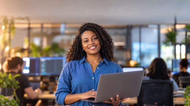 Desenvolvedora de software sorrindo, com camisa casual azul, segurando um notebook aberto, em um escritório de tecnologia moderno com monitores desfocados