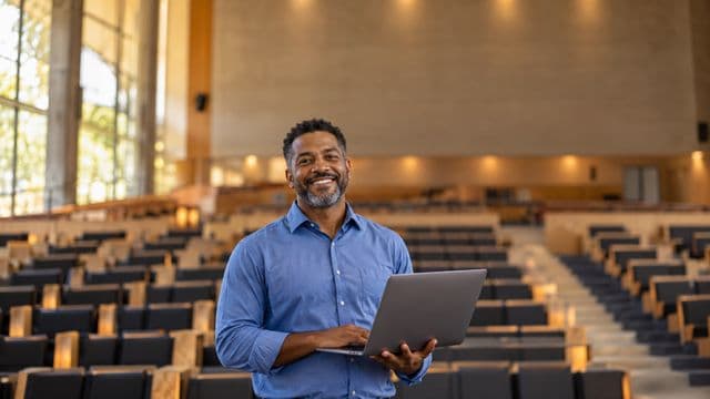 Professor universitário sorrindo, com camisa social azul, segurando um notebook, em um auditório universitário moderno