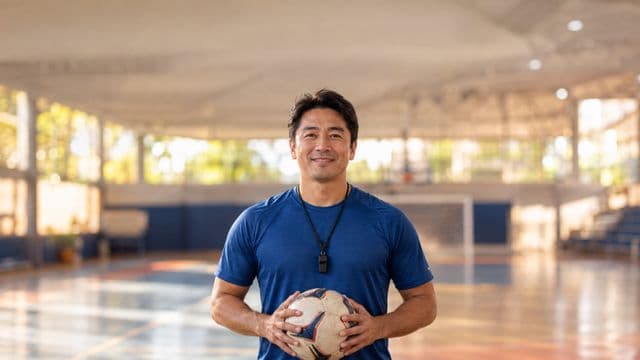 Profissional de educação física sorrindo, com camiseta esportiva azul e apito no pescoço, segurando uma bola, em uma quadra poliesportiva coberta e bem iluminada