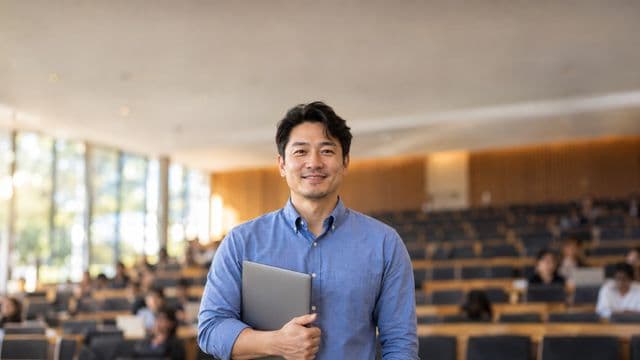 Professor universitário sorrindo, com camisa social azul, segurando um notebook, em um auditório universitário moderno