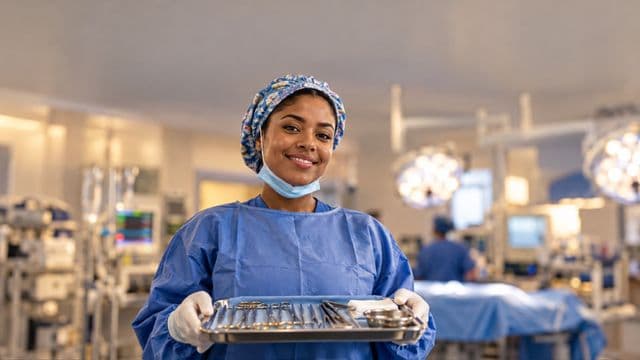 Instrumentadora cirúrgica sorrindo, com roupa cirúrgica azul, touca e máscara abaixada no queixo, segurando uma bandeja de instrumentos, em um centro cirúrgico moderno com focos de luz