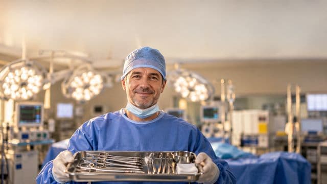 Instrumentador cirúrgico sorrindo, com roupa cirúrgica azul, touca e máscara abaixada no queixo, segurando uma bandeja de instrumentos, em um centro cirúrgico moderno com focos de luz