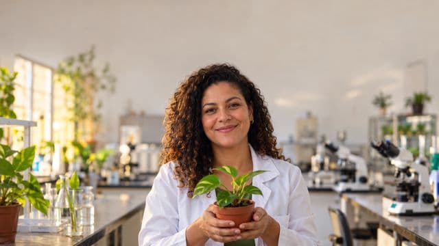Professora de biologia sorrindo, com jaleco branco, segurando uma planta em um vaso pequeno, em um laboratório de biologia com microscópios