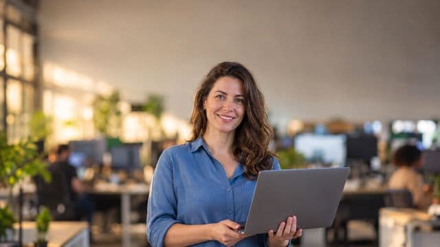 Desenvolvedora de software sorrindo, com camisa casual azul, segurando um notebook aberto, em um escritório de tecnologia moderno com monitores desfocados