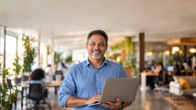 Empreendedor sorrindo, com camisa social azul com as mangas dobradas, segurando um notebook, em um espaço de coworking moderno e iluminado