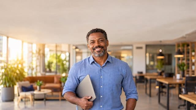 Empreendedor sorrindo, com camisa social azul com as mangas dobradas, segurando um notebook, em um espaço de coworking moderno e iluminado