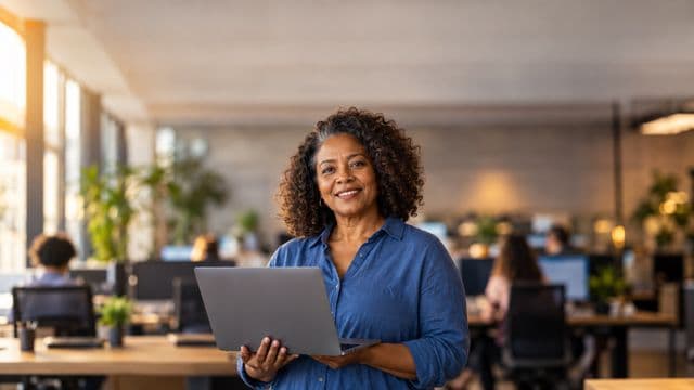 Profissional de tecnologia sorrindo, com camisa casual azul, segurando um notebook aberto, em um escritório de tecnologia moderno com monitores desfocados