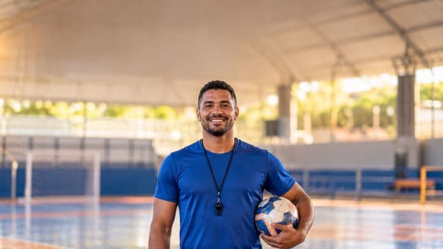 Profissional de educação física sorrindo, com camiseta esportiva azul e apito no pescoço, segurando uma bola, em uma quadra poliesportiva coberta e bem iluminada