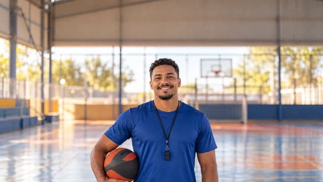 Profissional de educação física sorrindo, com camiseta esportiva azul e apito no pescoço, segurando uma bola, em uma quadra poliesportiva coberta