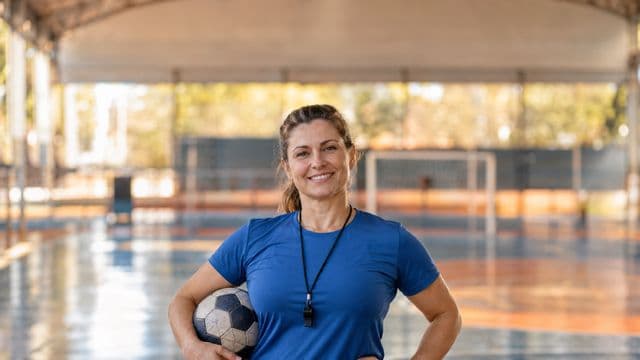 Profissional de educação física sorrindo, com camiseta esportiva azul e apito no pescoço, segurando uma bola, em uma quadra poliesportiva coberta e bem iluminada