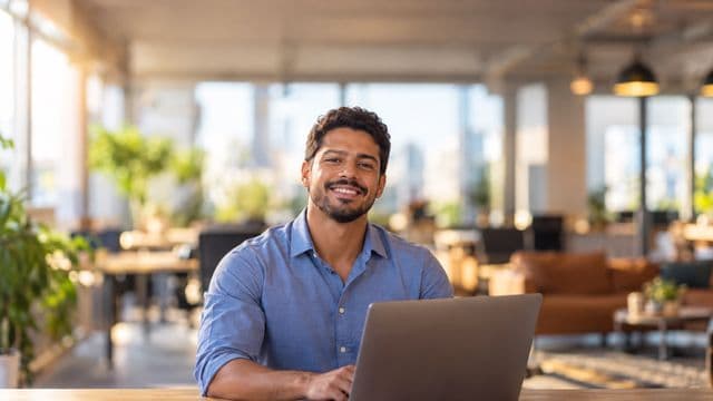 Empreendedor sorrindo, com camisa social azul com as mangas dobradas, segurando um notebook, em um espaço de coworking moderno e iluminado