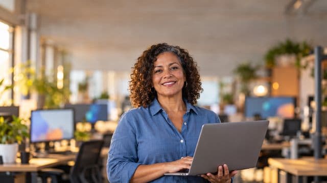 Desenvolvedora de software sorrindo, com camisa casual azul, segurando um notebook aberto, em um escritório de tecnologia moderno com monitores desfocados