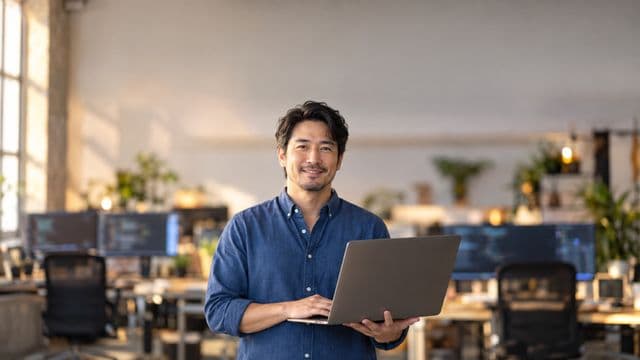 Desenvolvedor de software sorrindo, com camisa casual azul, segurando um notebook aberto, em um escritório de tecnologia moderno com monitores desfocados