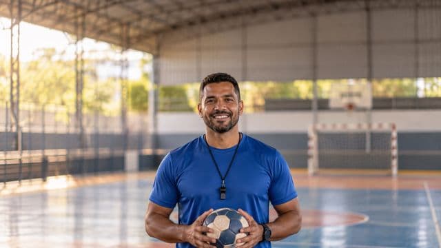 Profissional de educação física sorrindo, com camiseta esportiva azul e apito no pescoço, segurando uma bola, em uma quadra poliesportiva coberta e bem iluminada