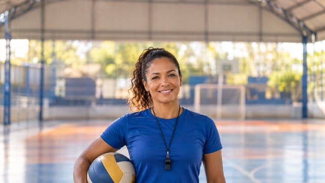 Profissional de educação física sorrindo, com camiseta esportiva azul e apito no pescoço, segurando uma bola, em uma quadra poliesportiva coberta e bem iluminada