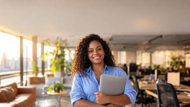 Empreendedora sorrindo, com camisa social azul com as mangas dobradas, segurando um notebook, em um espaço de coworking moderno e iluminado