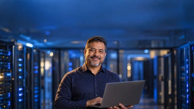 Especialista em segurança da informação sorrindo, com camisa social azul-escura, segurando um notebook, em uma sala de servidores com luzes azuis
