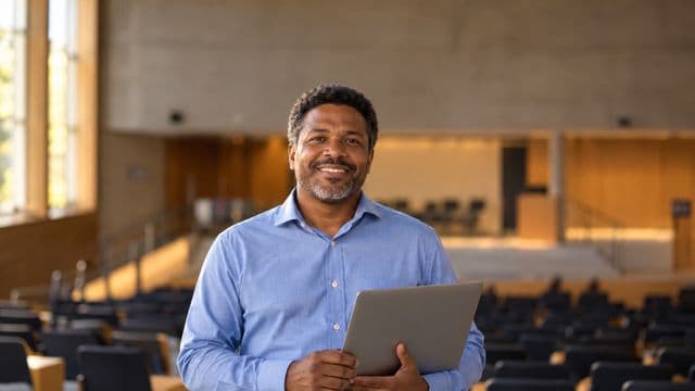 Professor universitário sorrindo, com camisa social azul, segurando um notebook, em um auditório universitário moderno
