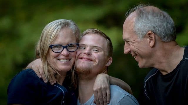 Pai e mãe abraçam o filho jovem com síndrome de Down, os três sorrindo, em área verde ao ar livre