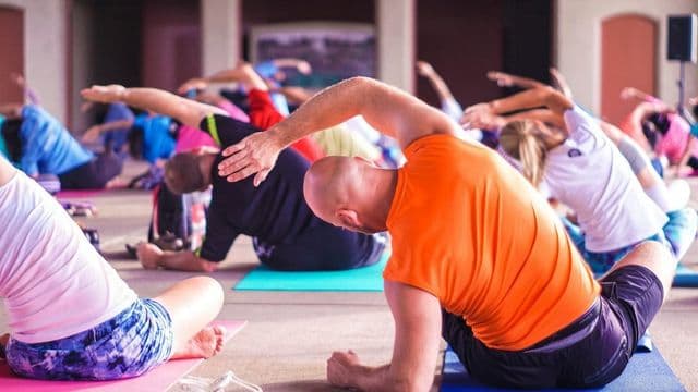 Aula coletiva de alongamento: pessoas de várias idades se exercitam sentadas em colchonetes coloridos, com um homem de camiseta laranja em primeiro plano