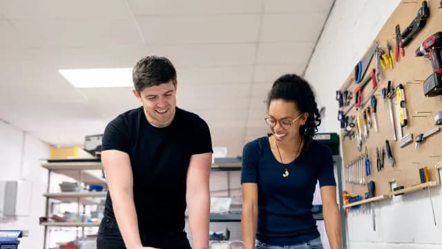 Engenheiro e engenheira sorrindo em uma bancada de laboratório com ferramentas na parede