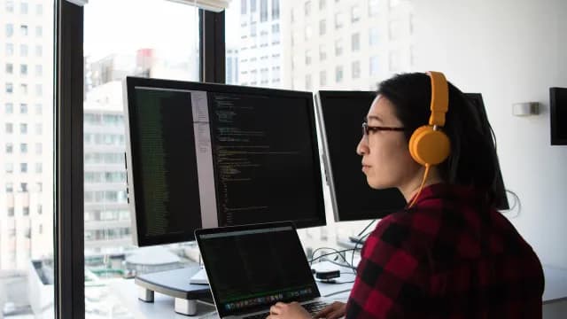 Engenheira de software com fones de ouvido programando em dois monitores, em um escritório com vista para a cidade