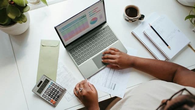 Gestora analisa planilha de orçamento no notebook com calculadora, recibos e caderno de anotações sobre a mesa