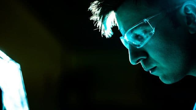 Perito em computação forense concentrado examina a tela do computador em laboratório escuro, com o rosto iluminado pela luz azul do monitor