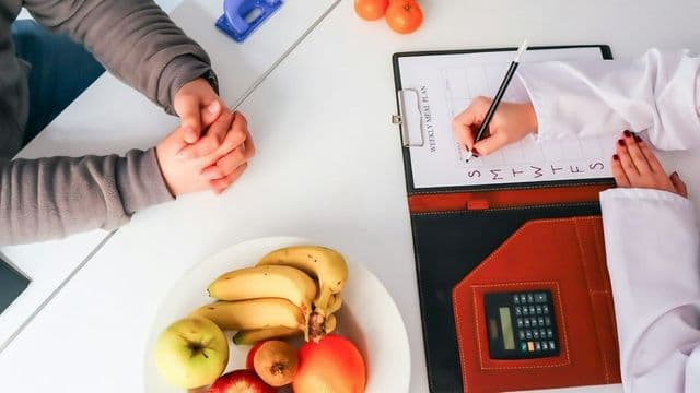 nutricionista preenche plano de refeições semanal ao lado de paciente, com frutas frescas na mesa