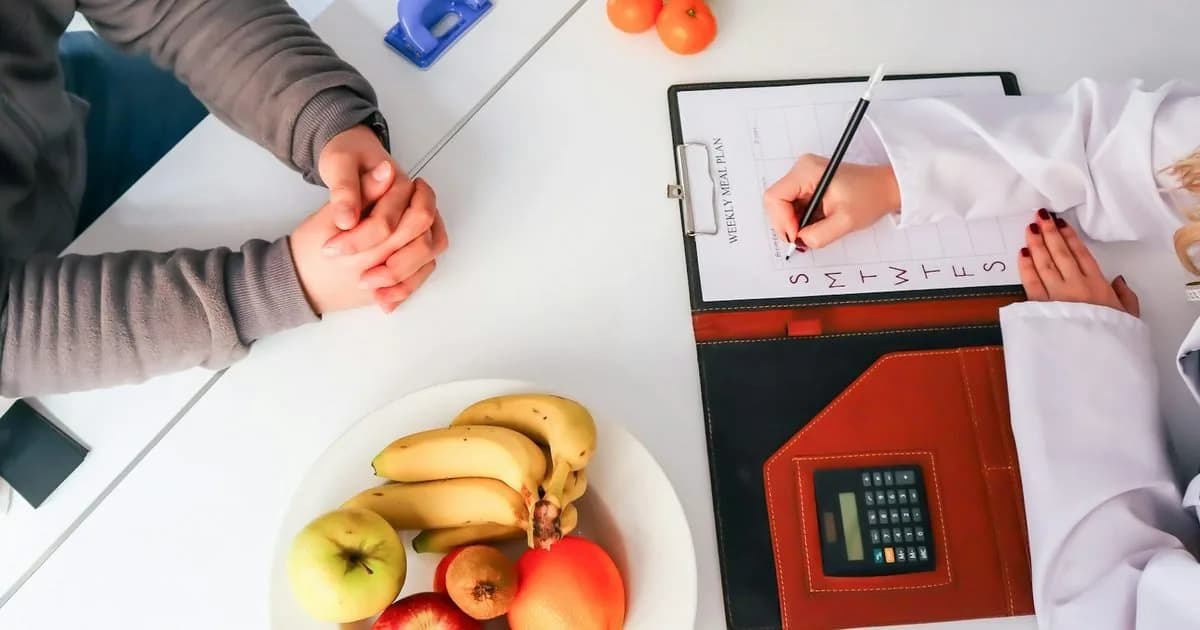 nutricionista preenche plano de refeições semanal ao lado de paciente, com frutas frescas na mesa