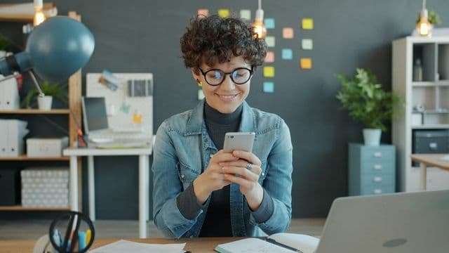 Mulher sorri enquanto mexe no celular em um escritório criativo de comunicação, com post-its coloridos ao fundo