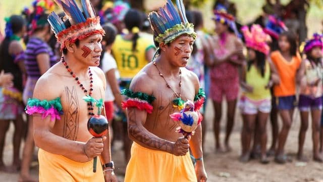 Homens indígenas com cocar de penas conduzem celebração tradicional na aldeia, com crianças e jovens da comunidade ao fundo