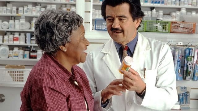 Farmacêutico de jaleco branco orienta uma cliente no balcão de uma farmácia, com prateleiras de medicamentos ao fundo