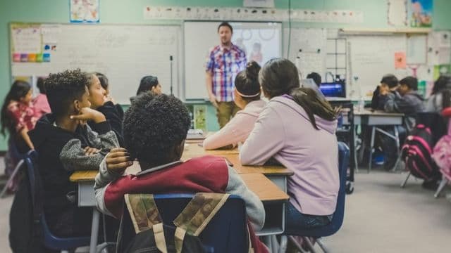 Professor em sala de aula conduzindo os alunos, com mochilas apoiadas nas carteiras, representando a gestão escolar no Brasil