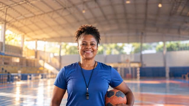 Profissional de educação física sorrindo, com camiseta esportiva azul e apito no pescoço, segurando uma bola, em uma quadra poliesportiva coberta e bem iluminada