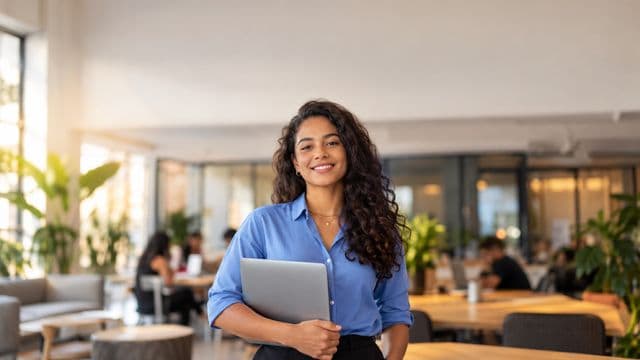 Empreendedora sorrindo, com camisa social azul com as mangas dobradas, segurando um notebook, em um espaço de coworking moderno e iluminado